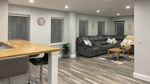 A modern living room with a grey sofa, a wooden coffee table, and a dining area with a wooden table and bar stools. - Spacious 5Br Home - Country View - Close to Galway (Tuam)