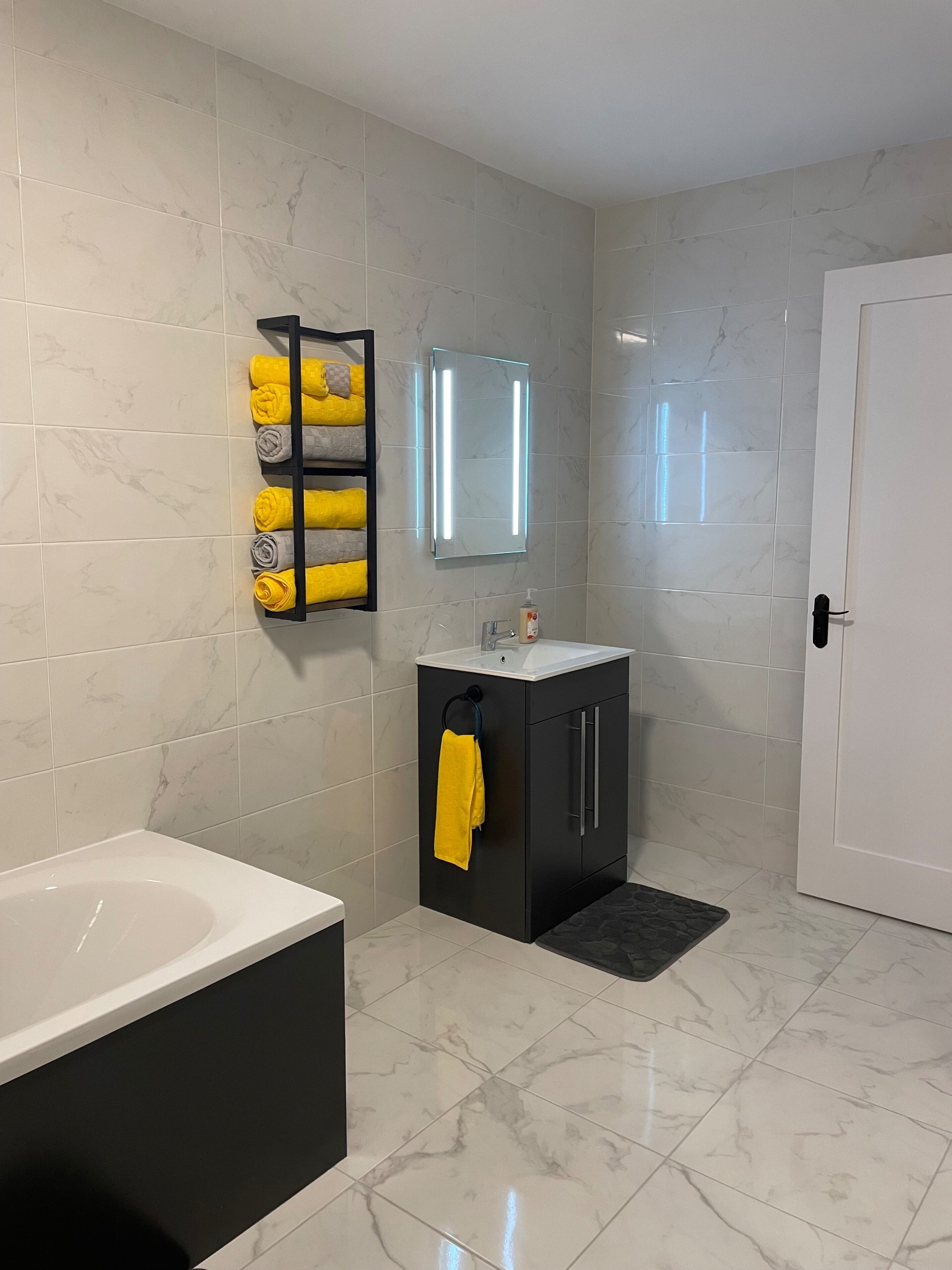 Un baño con una bañera blanca, un tocador negro con un espejo y un perchero con toallas amarillas.