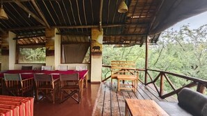 Uma área de refeições ao ar livre coberta, com mesas e cadeiras de madeira, um deck de madeira e vista para a floresta circundante.