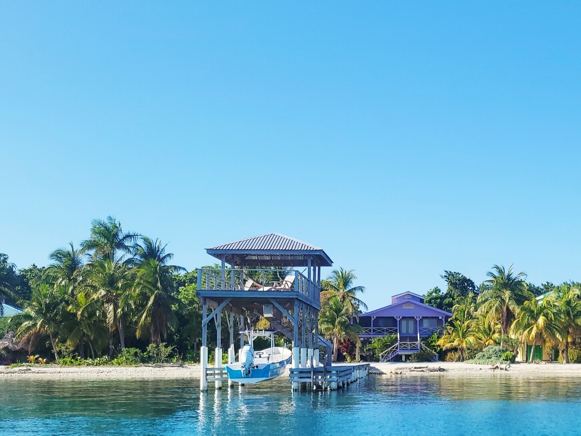 A boat docked at a pier with a covered structure, surrounded by palm trees and a beach.