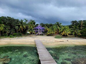Una casa de playa con una terraza de madera que conduce a la entrada, rodeada de palmeras y una vista despejada del océano.