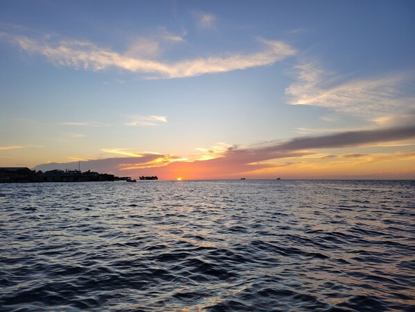 A coastal view with a sunset over the sea, buildings along the shore, and a calm water surface.