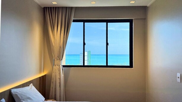 Ein Hotelzimmer mit großem Fenster, das Meer und Gebäude zeigt, einem Bett mit weißen Leinen und einem Vorhang.