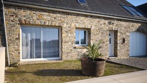 Une maison en pierre avec de grandes fenêtres, une porte et un petit jardin agrémenté d’une plante en pot.