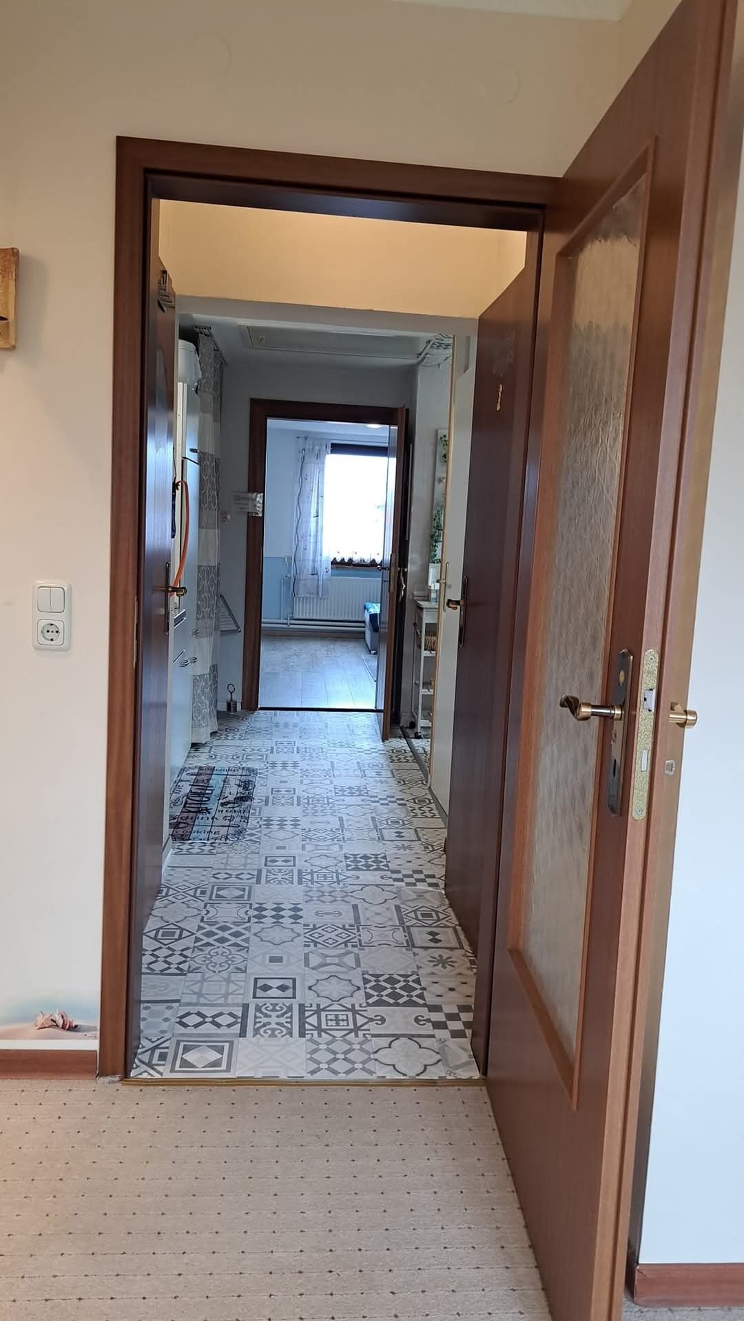 A hallway with patterned carpet leading to a room with a window and a door.