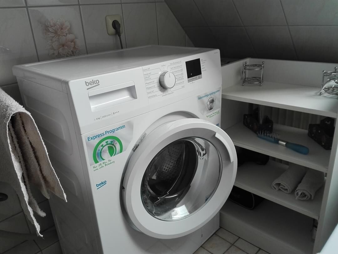 Eine Waschmaschine mit Frontlader, ein Fach mit gefalteten Handtüchern und eine Steckdose an der Wand.