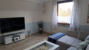 A living room with a flat-screen TV, a sofa, a coffee table with a glass top, a potted plant, and a window with curtains.