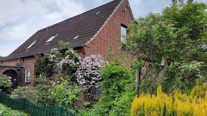 A brick house with a sloped roof, a green fence, and various plants and trees.