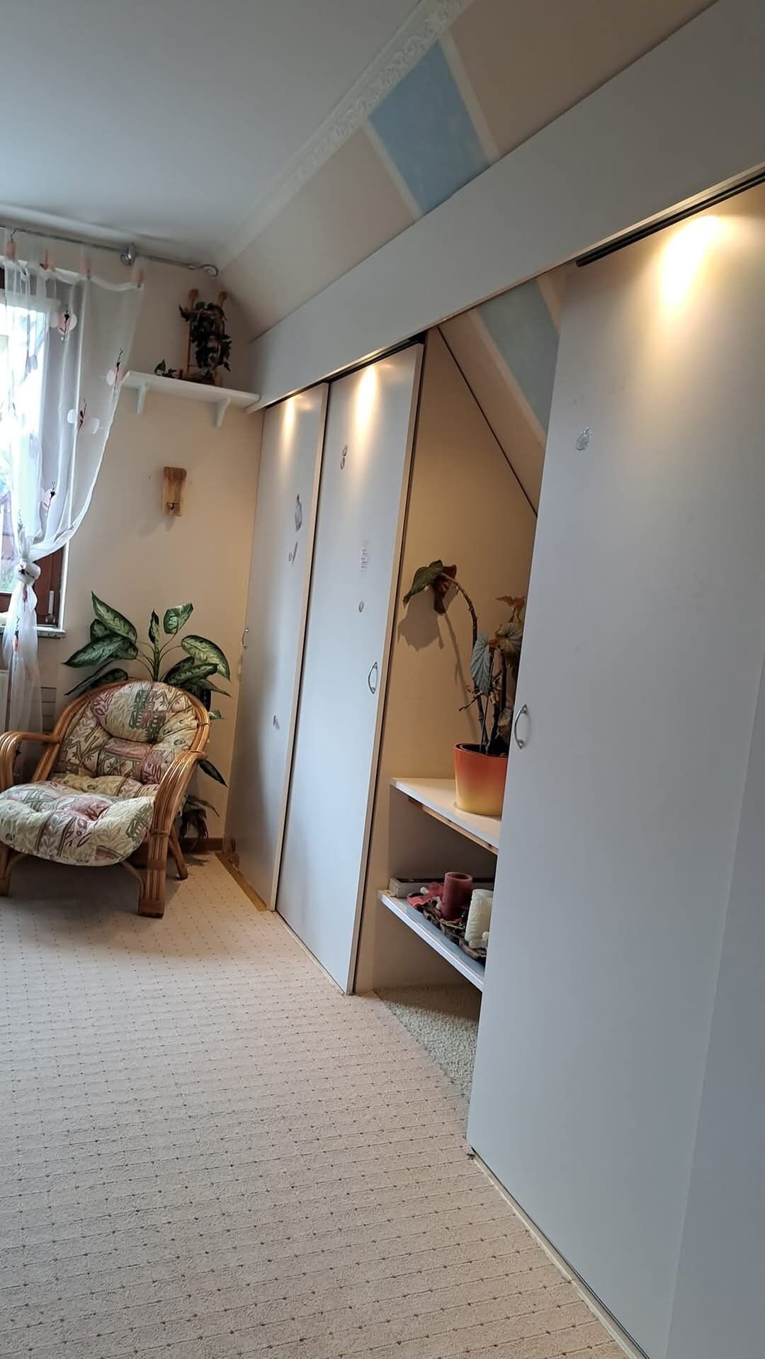 Um quarto com uma cadeira estampada, uma planta em vaso, um guarda-roupa e uma estante com objetos.