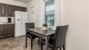 Een kleine keuken met donkerhouten kastjes, een witte koelkast en een koffiezetapparaat op het aanrecht. Er staat een eettafel met twee stoelen en een kleine potplant op de vensterbank.
