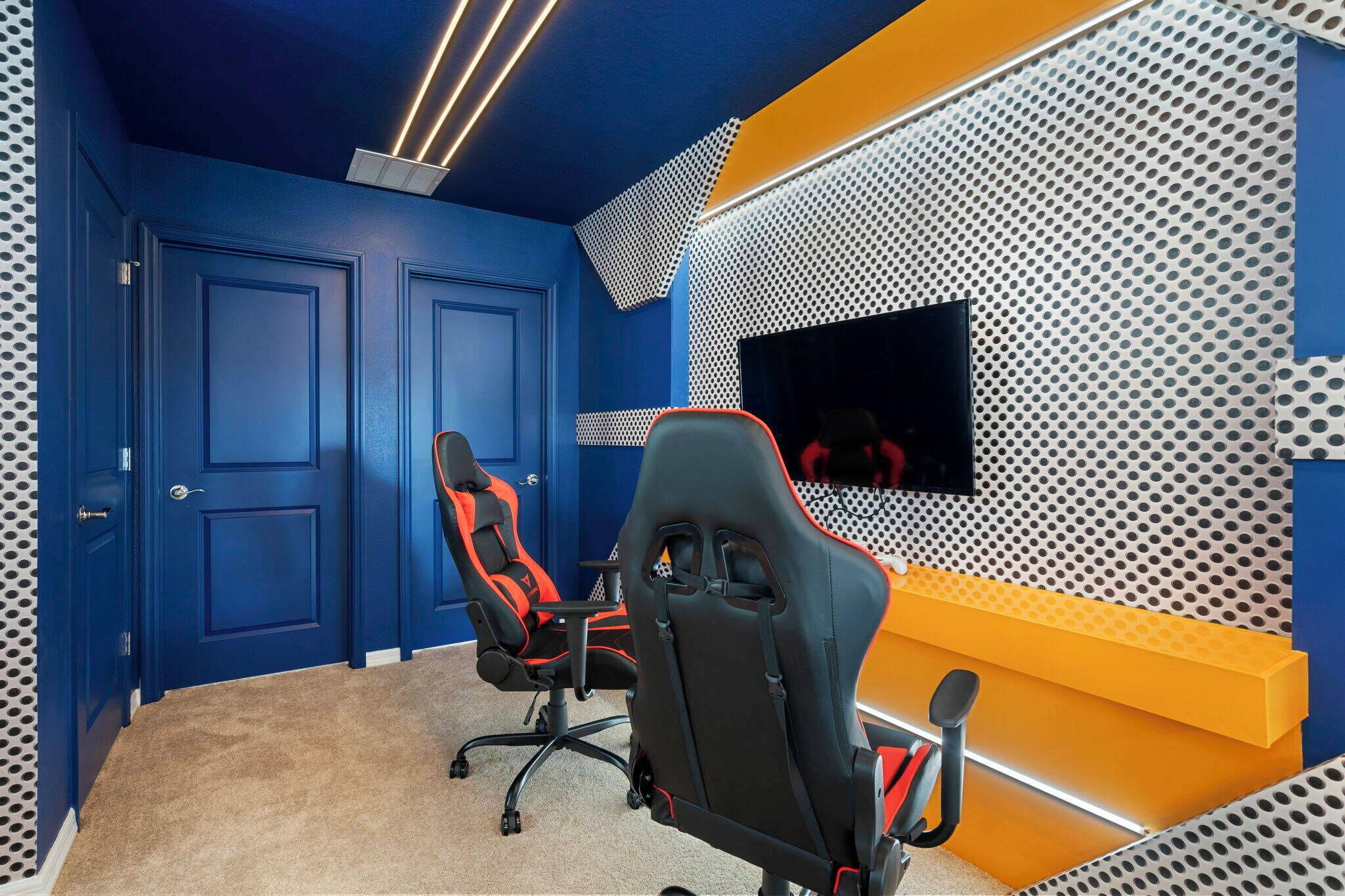 A room with two doors, a gaming chair, a TV mounted on a perforated wall, and a yellow bench.