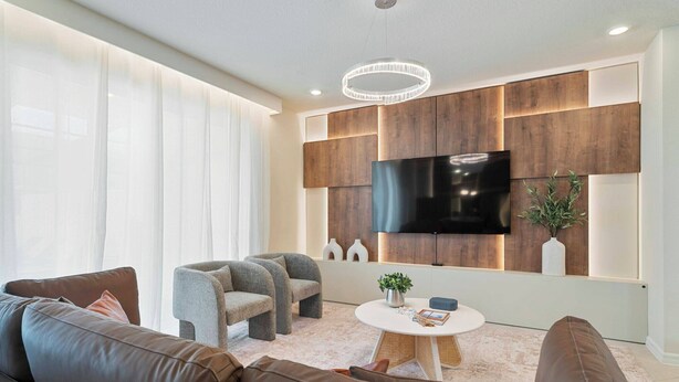 A modern living room with a large flat-screen TV mounted on a wooden panel wall, a round coffee table with a plant, and a seating area with leather sofas and armchairs. - Stunning Pool Home just minutes from Disney 12 (Clermont)