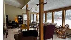 Una sala de estar acogedora con un sofá marrón, un sillón amarillo y una mecedora. Amplias ventanas ofrecen vistas a un paisaje nevado.