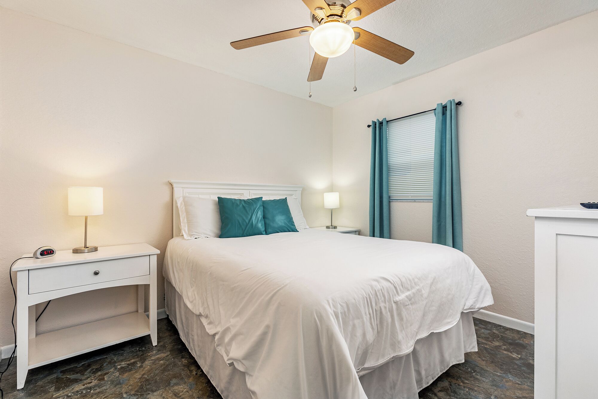 Une chambre à coucher comprenant un lit, deux tables de chevet, une commode, un ventilateur de plafond et une fenêtre avec des rideaux.