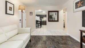 Un salon avec un canapé blanc, un coin repas avec une table et des chaises, une cuisine à l’arrière-plan et un couloir menant à une autre pièce.