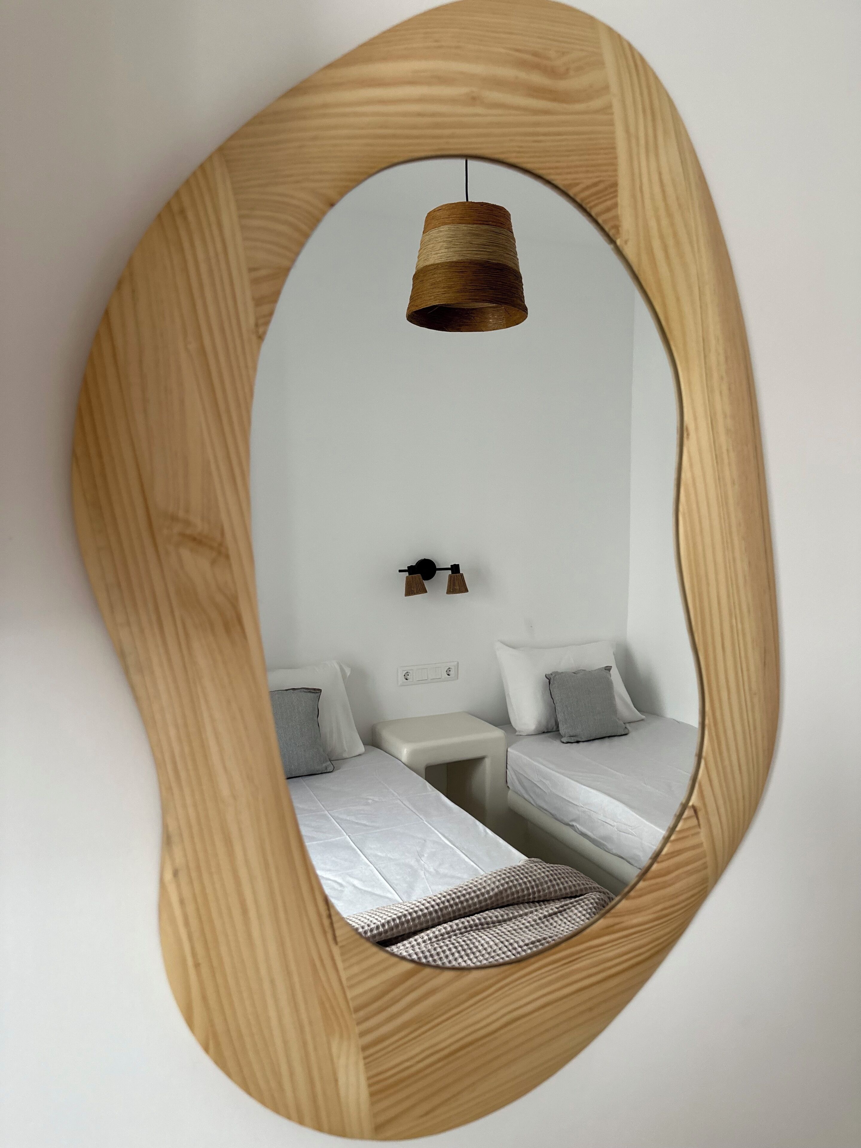 A room with a wooden-framed mirror, a hanging light fixture, two beds with pillows, a small bedside table, and a power outlet on the wall.