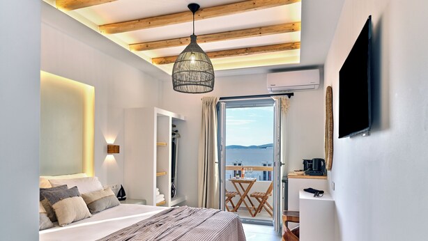 A bedroom with a bed, a hanging light fixture, a TV, and a view of the sea. - Marjay (Paros)