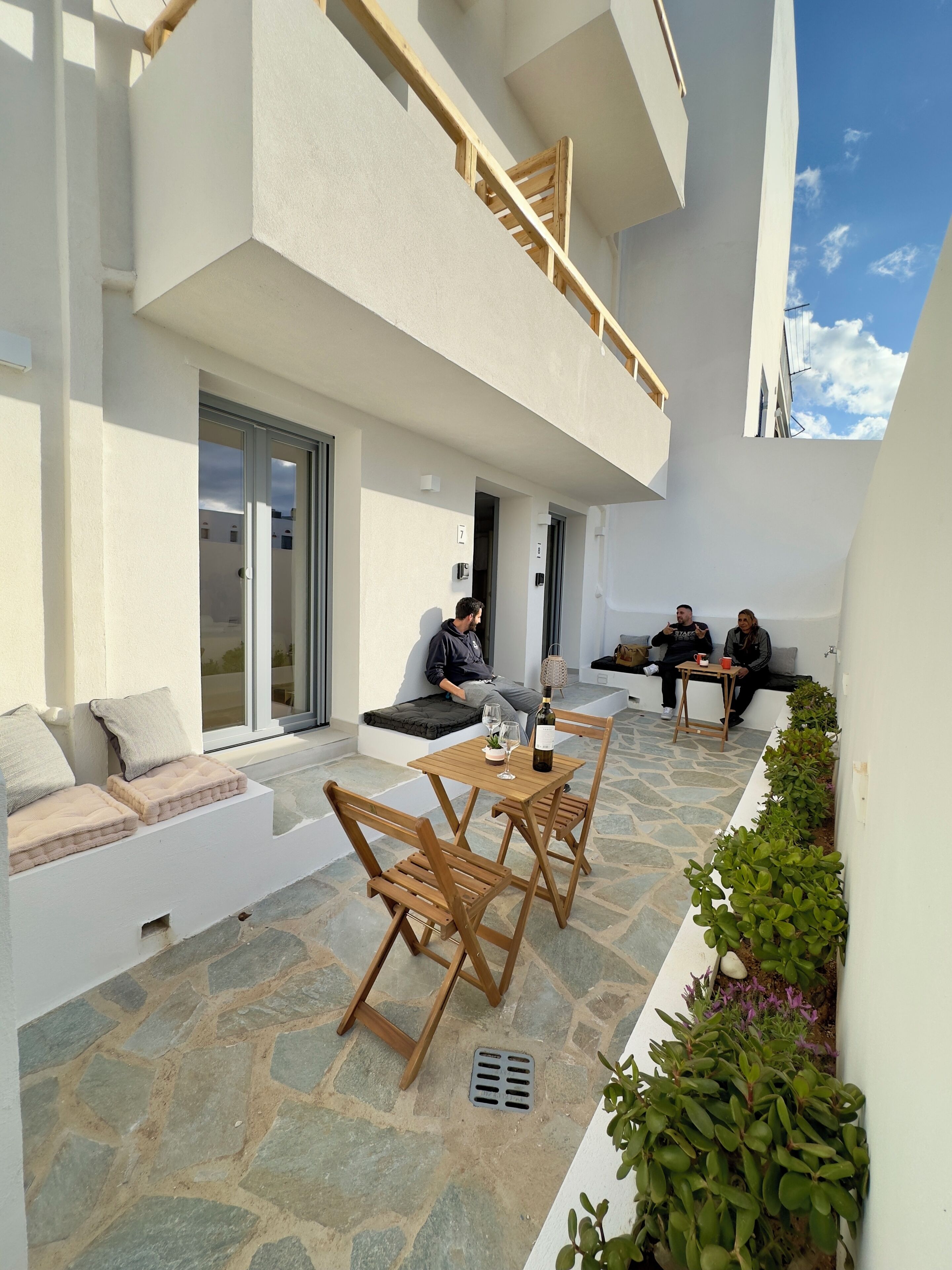 A modern outdoor seating area with stone flooring, wooden furniture, and a small table with a bottle and glasses.