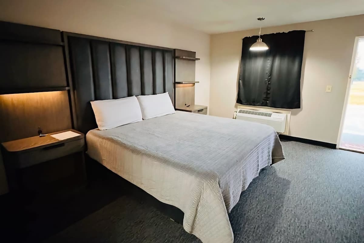 A modern hotel room with a large bed, a bedside table, a nightstand, a wall-mounted shelf, and a window with curtains.