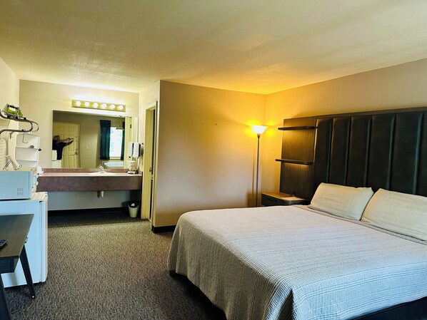 A hotel room with a bed, a desk, a chair, a mirror, a sink, and a nightstand.
