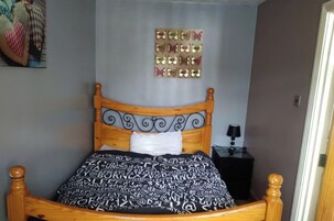 A bed with a patterned bedspread, a wooden headboard with decorative scrollwork, a wall-mounted mirror, and a small bedside table with a lamp.