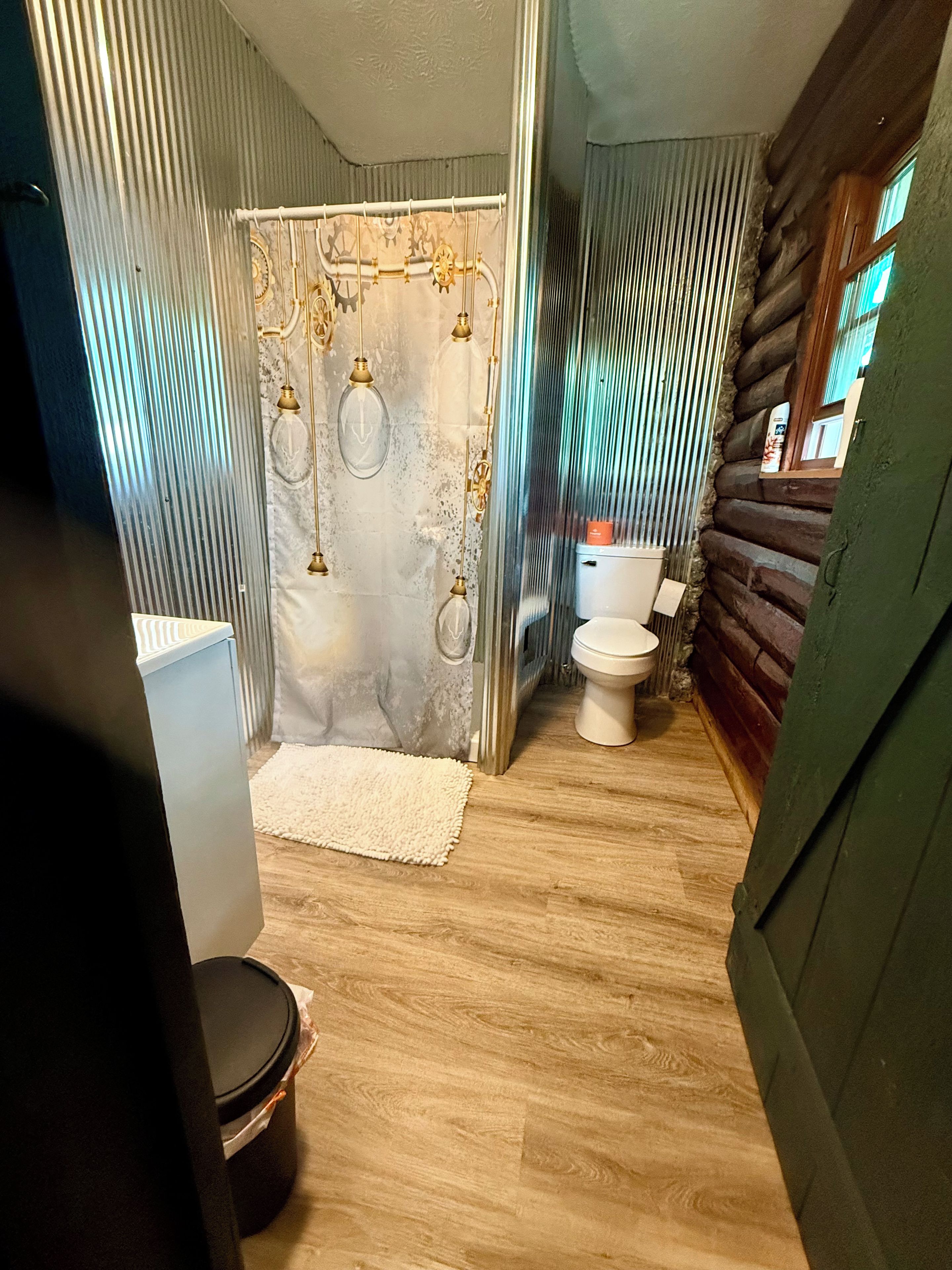 A bathroom with a wooden floor, a white toilet, a shower curtain with hanging lights, and a white rug.