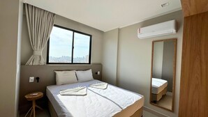 A hotel room with a large bed, a small bedside table, a window with curtains, an air conditioning unit, and a mirror. - Studio Design 3 Apt 803 (Maceio)