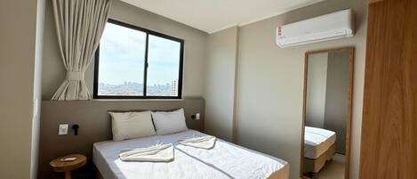 A hotel room with a large bed, a small bedside table, a window with curtains, an air conditioning unit, and a mirror.