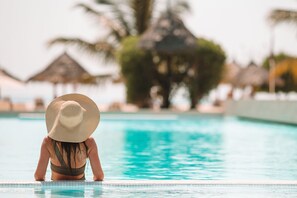 A person in a swimming pool with a large hat, surrounded by palm trees and thatched-roof structures.