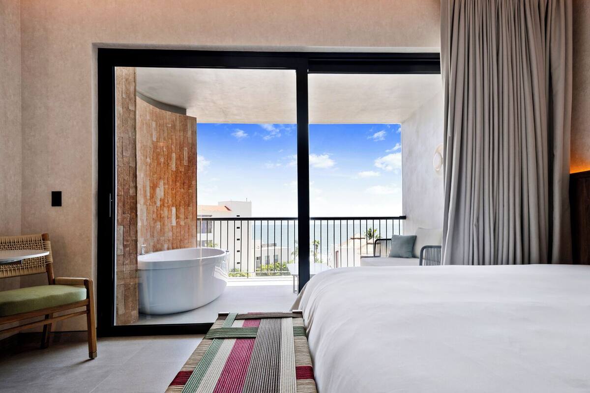 Ein modernes Hotelzimmer mit einem großen Bett, Blick auf den Strand, einer Badewanne und einem Balkon mit einem Sessel.