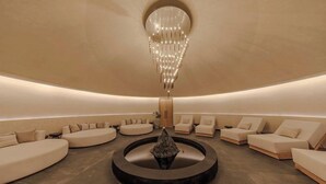 Egy modern wellness-szoba, melyben kör alakú ülősarok, központi vízfelület és egy díszes csillár található.