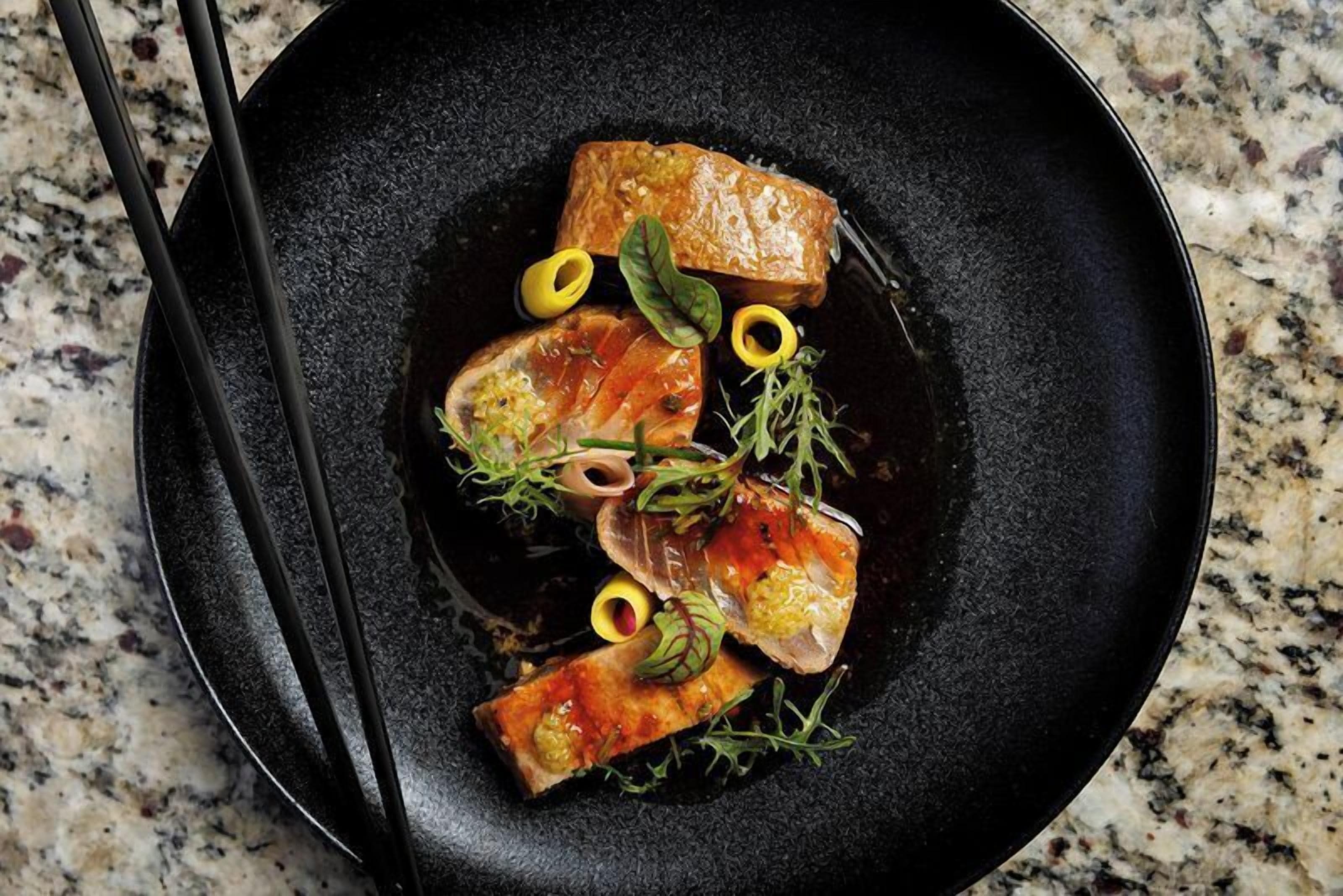 Un plato negro con comida, incluyendo salmón y verduras, sobre una encimera de granito.