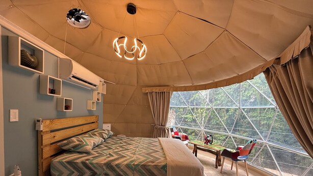 Un dormitorio con una cama grande, cabecera de madera, una lámpara de techo, una ventana con cortinas y vistas a árboles.