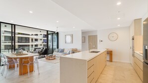Una cocina moderna con una isla grande, un comedor con sillas transparentes y una sala de estar con sofá y mesa de centro.