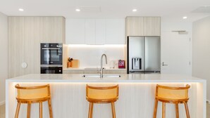 Una cocina moderna con una isla central, electrodomésticos de acero inoxidable y tres taburetes de mimbre.