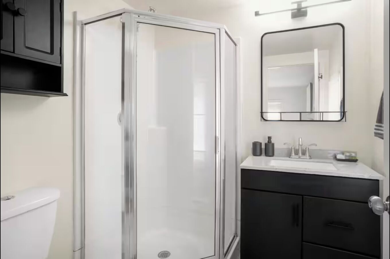 Un baño con cabina de ducha de vidrio, un lavamanos blanco con un espejo encima y un mueble negro debajo.
