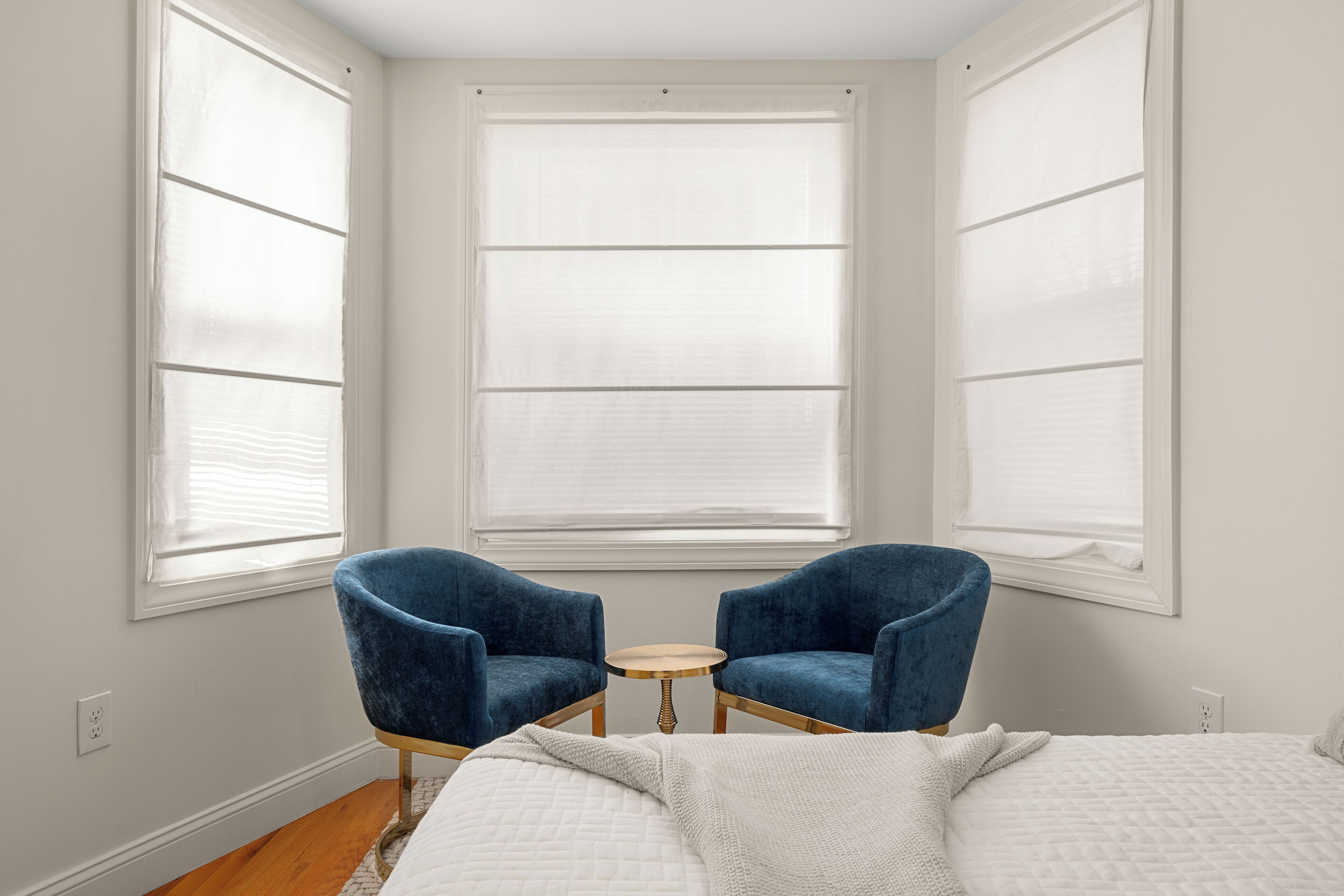 A bedroom with a bed, two blue armchairs, a small round table, and a large window with blinds.
