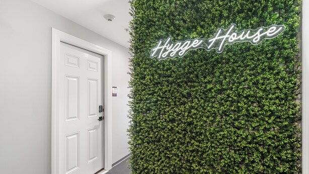 A hallway with a white door and a wall covered in green foliage with the text "Hygg House" on it. - Hygge House Suites (Newburyport)