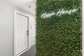 Ein Flur mit einer weißen Tür und einer Wand, die mit grünem Blattwerk bedeckt ist. Auf der Wand ist der Schriftzug 'Hygg House' zu sehen.