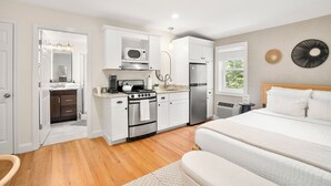 A modern kitchen with white cabinets, a stove, and a refrigerator. There is a wooden floor and a bed with white bedding in the adjacent room. - Hygge House Suites (Newburyport)