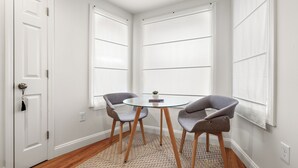 A corner room with a glass table, two grey chairs, a small plant, and a white door. - Hygge House Suites (Newburyport)