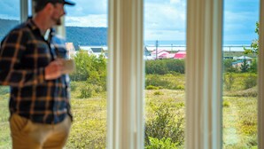 Une personne se tient près d’une fenêtre, d’où l’on peut admirer un paysage côtier comprenant des bâtiments et de la végétation.