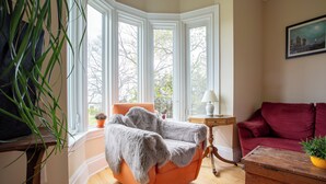 Une pièce avec une fenêtre d’angle, un fauteuil recouvert d’une peau de bête, un canapé rouge et une commode en bois.