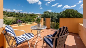 A balcony with two striped chairs and a small table, overlooking a scenic landscape with trees and buildings. - Villas del Golf townhouse 27 (Manilva)