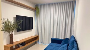 A modern living room with a blue sofa, a mounted TV, a wooden shelf with a plant, and a window with curtains. - Studio Design 3 Apt 902 (Maceio)