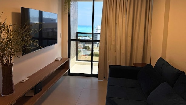 A modern living room with a flat-screen TV, a dark sofa, a wooden bench, and a potted plant. - Studio Design 3 Apt 902 (Maceio)