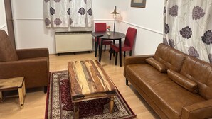 Ένα σαλόνι με έναν καφέ δερμάτινο καναπέ, ένα ξύλινο τραπεζάκι σαλονιού, μια κόκκινη καρέκλα, ένα μικρό τραπέζι και ένα καλοριφέρ.