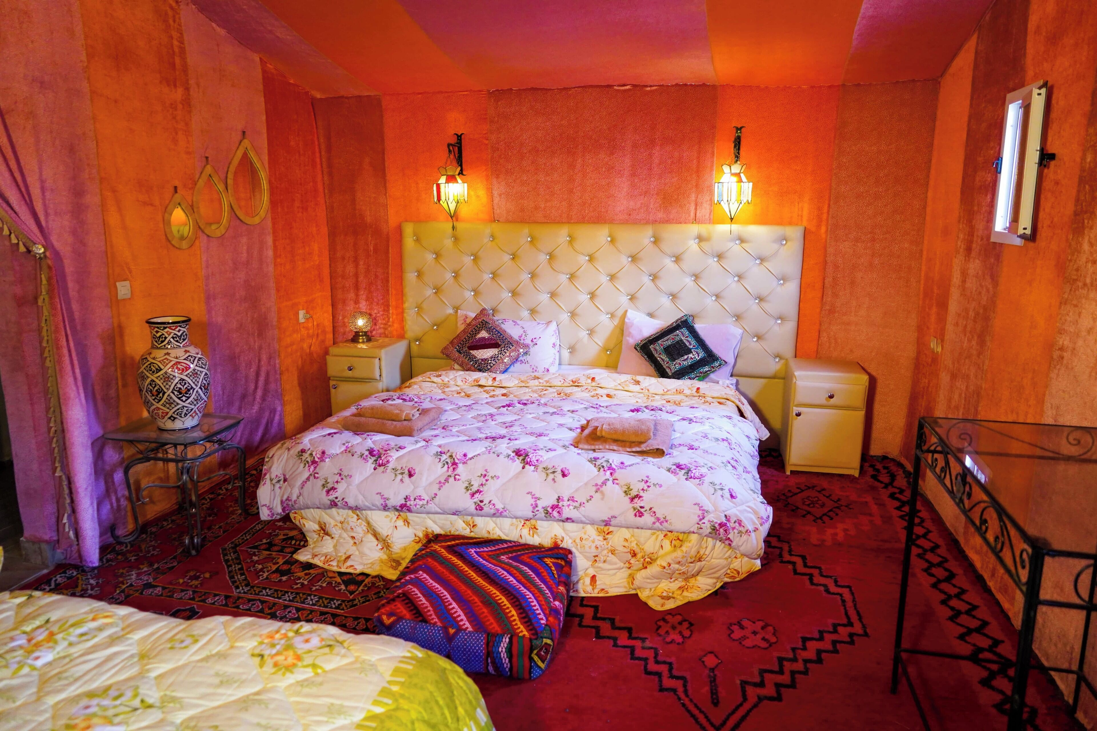 A bedroom with a tufted headboard, floral bedding, two bedside lamps, a nightstand, a decorative vase, and a patterned rug.