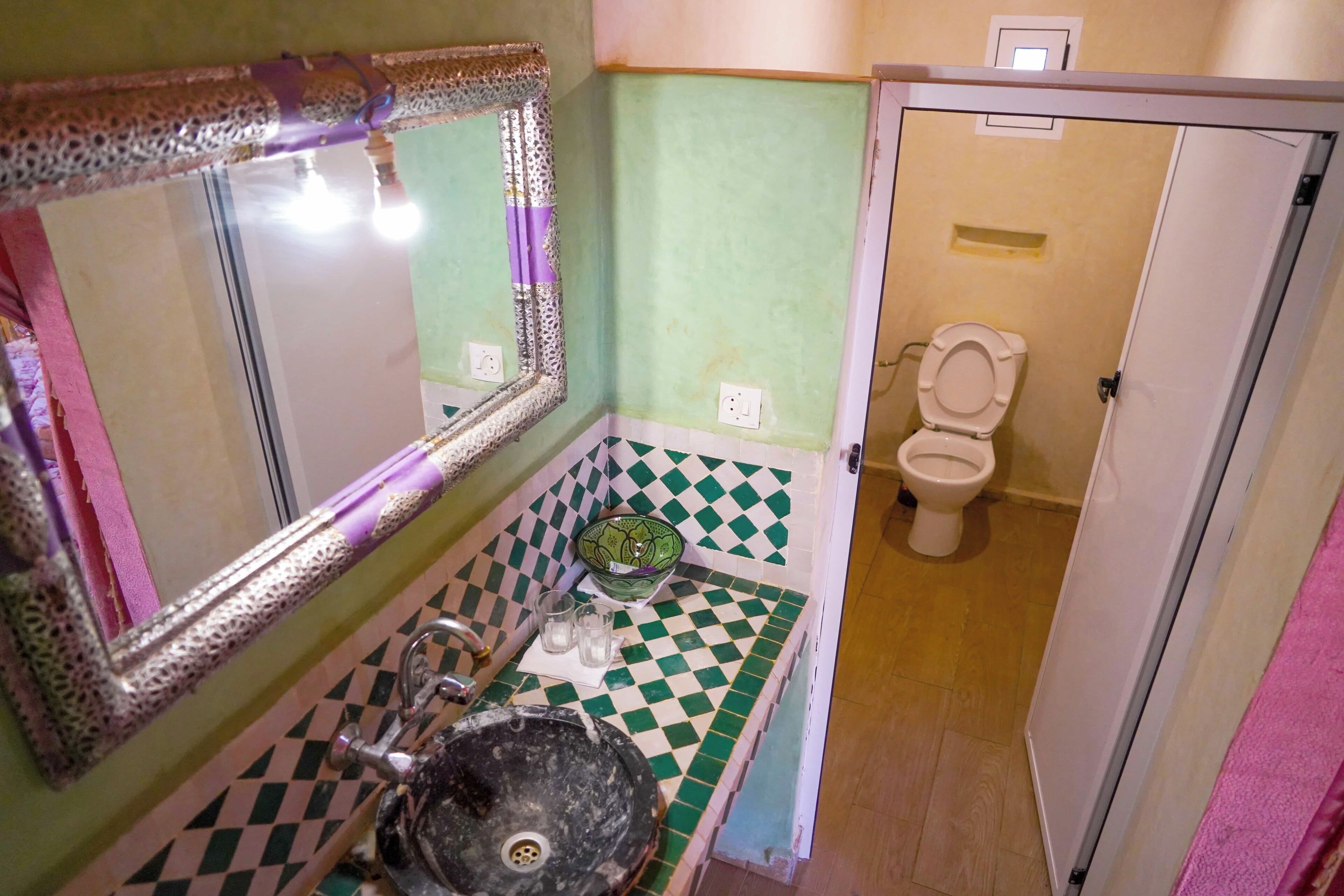 A bathroom with a green and white checkered floor, a sink with a decorative mirror, a green bowl, and a toilet.