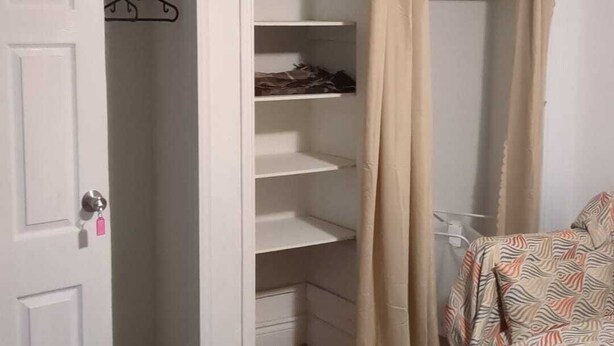 A room with a white door, a closet with shelves, a bed with a patterned bedspread, and a black rug. - Basement Unit (Richmond Hill)